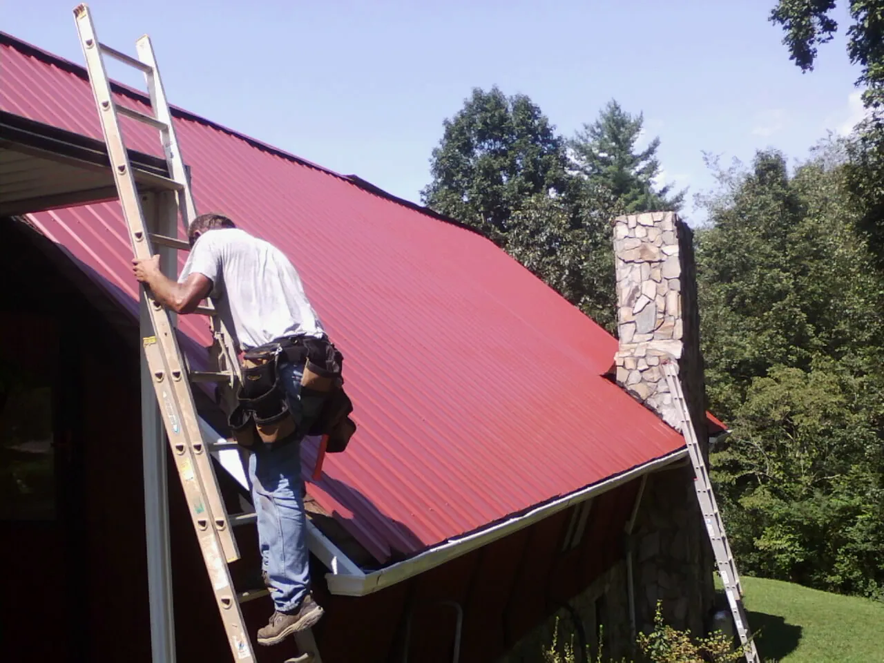 Roofer on a ladder beside a red metal roof and stone chimney — Soffit & Fascia Repair by Roofing Brothers in Andrews